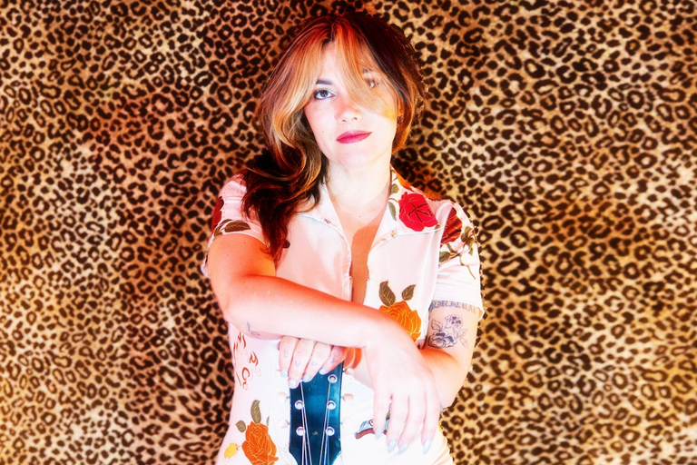 Australian singer-songwriter Ella Hooper leaning on a guitar headstock in a floral top against a leopard print backdrop.
